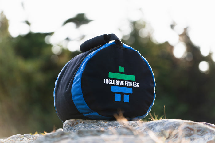 Big Land Throwing Sandbag with its grab handle, resting on a rock outdoors, black and blue with the Inclusive Fitness logo