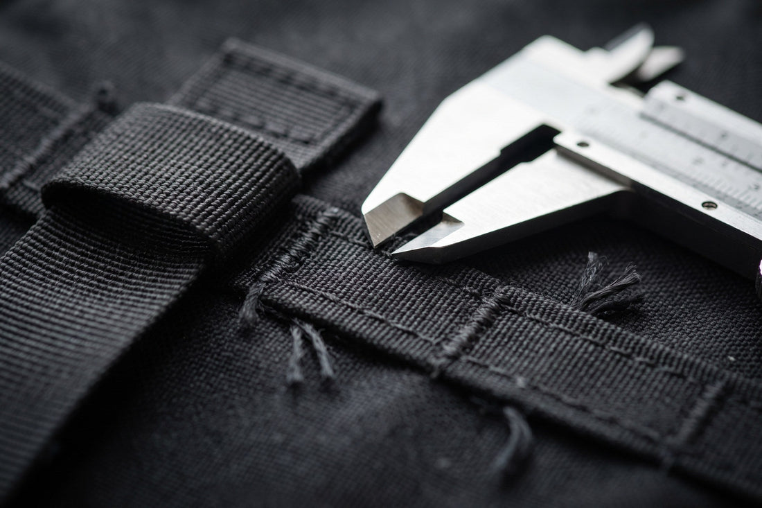 Close-up of heavy bar-tack stitching on black webbing beside a steel caliper