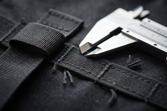 Close-up of heavy bar-tack stitching on black webbing beside a steel caliper