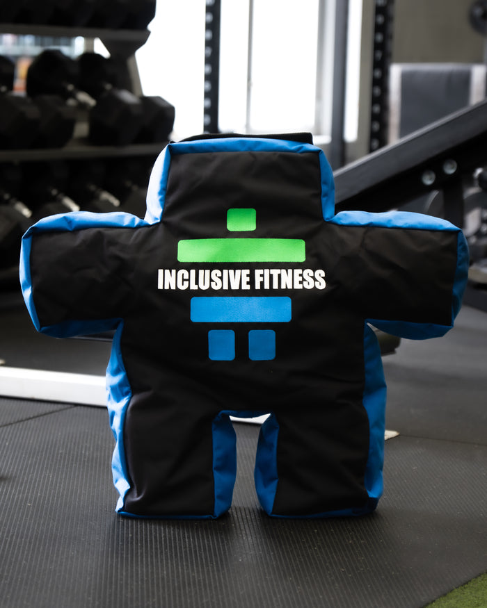 The Inukshuk Sandbag, a human shaped strongman sandbag in black and blue with the Inclusive Fitness logo, standing in a gym